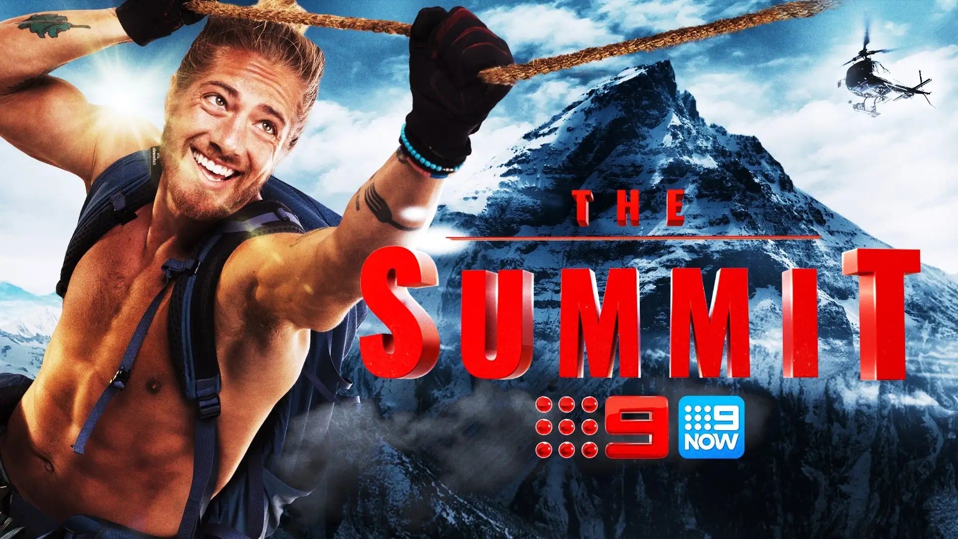 A muscular man with a beard and tattoos, wearing black gloves and a blue backpack strap over his shoulder, is gripping a thick rope while smiling against a backdrop of snow-capped mountains.