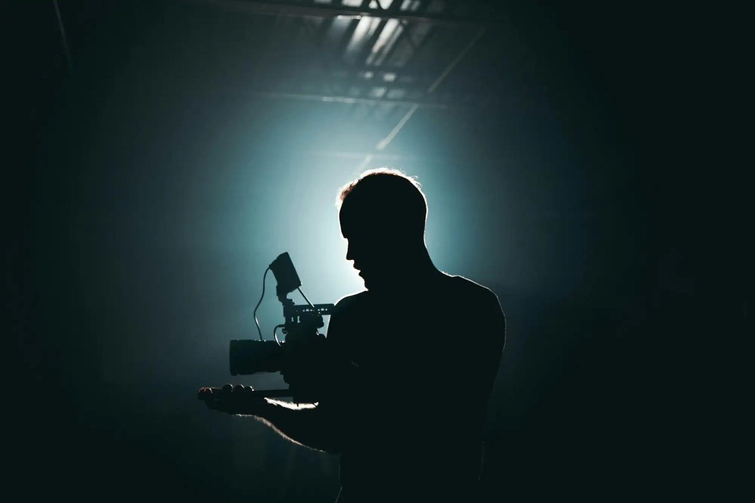 Silhouetted filmmaker holding a professional cinema camera with attached lighting rig against dramatic backlighting.