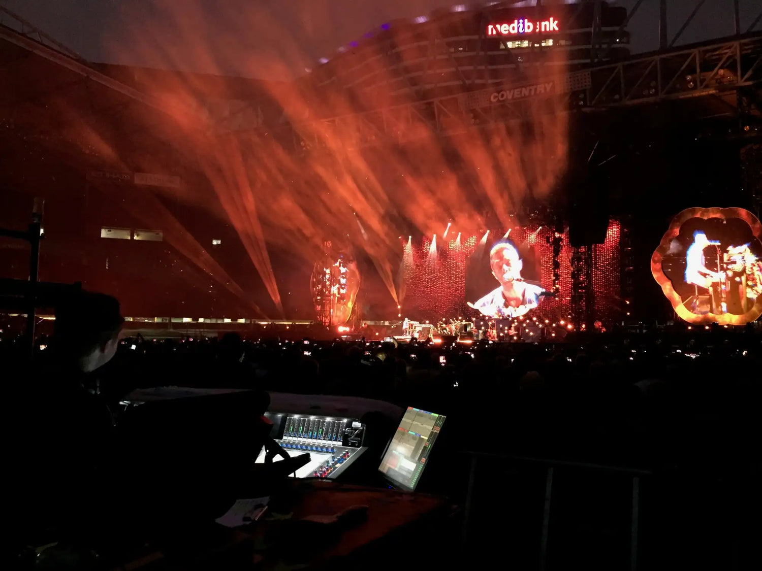 A vibrant red stage illuminated by sweeping laser lights, featuring a large central screen displaying the performer and flanked by dynamic visual effects.